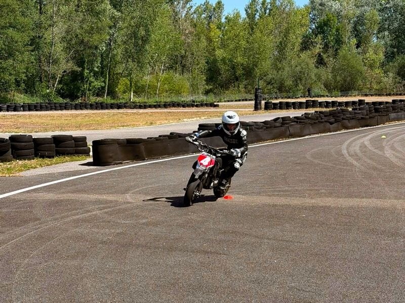Pilote en combinaison noire et casque blanc, en virage serré sur une moto noire sur circuit avec pneus en arrière-plan.