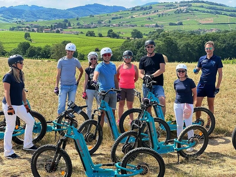 Groupe de sept personnes avec trottinettes électriques posant dans un champ verdoyant sous un ciel clair.