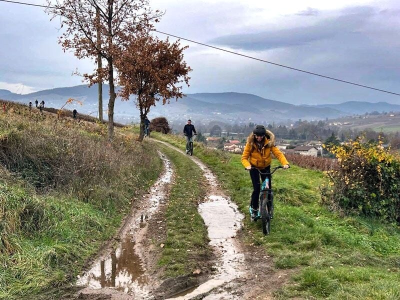 Personnes en trottinettes électriques sur un sentier boueux avec des collines et un village en arrière-plan.