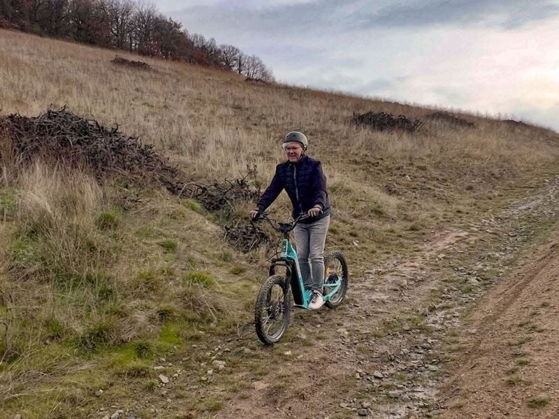 Femme souriante en trottinette électrique descendant un chemin de terre dans un paysage de collines sèches.