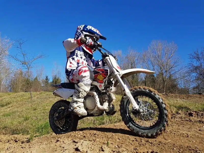 Jeune pilote en tenue complète sur une moto tout-terrain blanche dans un terrain boueux et herbeux sous un ciel bleu.