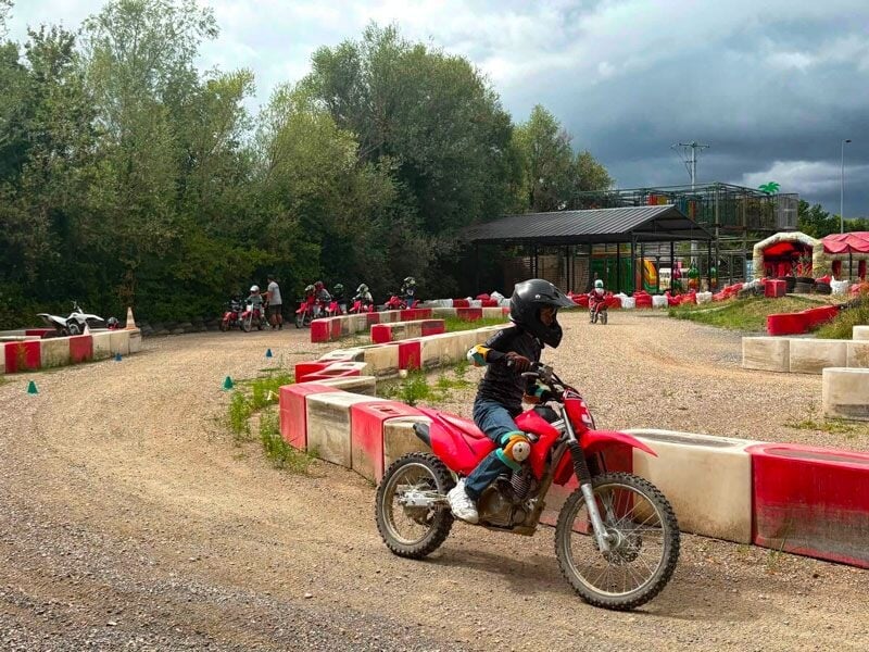 Enfant en casque noir sur une moto rouge sur une piste de karting avec barrières rouges et blanches et arbres en arrière-plan.