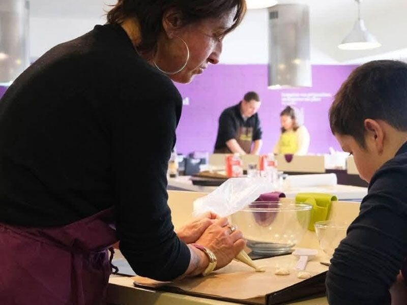 Une femme en tablier violet montre une technique de cuisine à un garçon dans une salle avec plusieurs participants.
