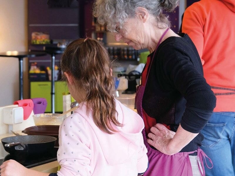 Une femme âgée en tablier violet guide une jeune fille en sweat rose lors d'un atelier de cuisine convivial.