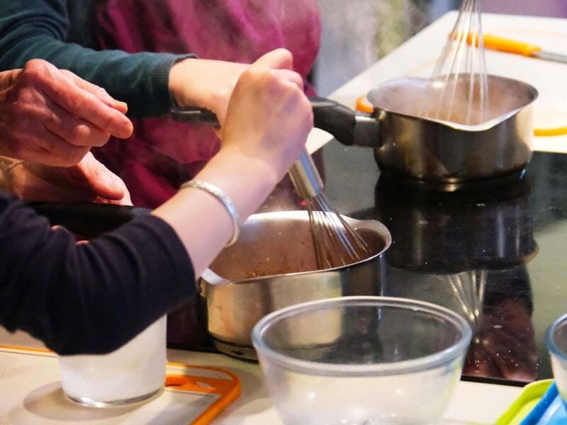 Des mains d'adultes et d'enfants fouettent un mélange dans une casserole sur une plaque de cuisson lors d'un cours.