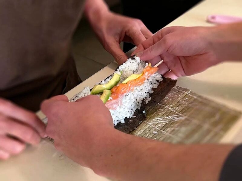 Deux personnes préparent un rouleau de sushi avec du riz, avocat et saumon sur une natte en bambou.