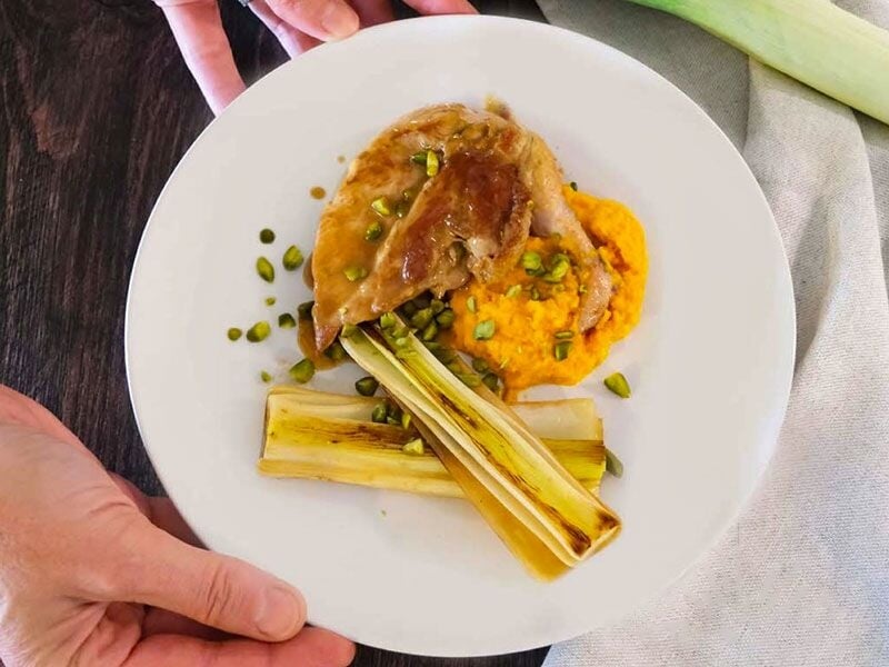 Un plat gastronomique avec viande, purée orange, poireaux et graines présenté sur une assiette blanche.