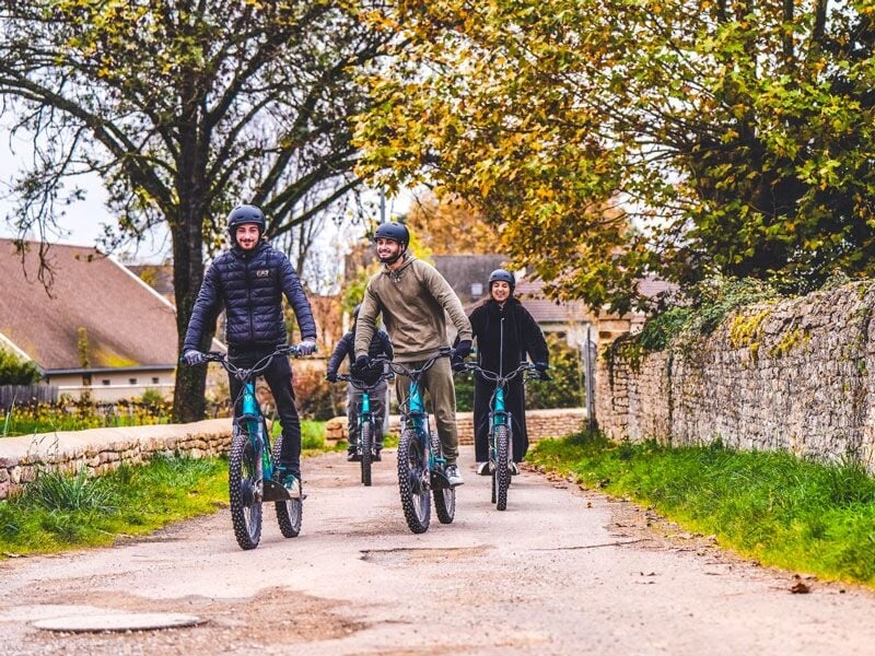 Quatre cyclistes portant des casques roulent sur une route bordée d'arbres et de murs en pierre en automne.
