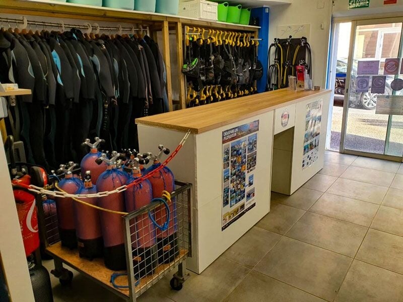Intérieur d'un centre de plongée avec combinaisons suspendues et bouteilles d'air comprimé alignées au sol.