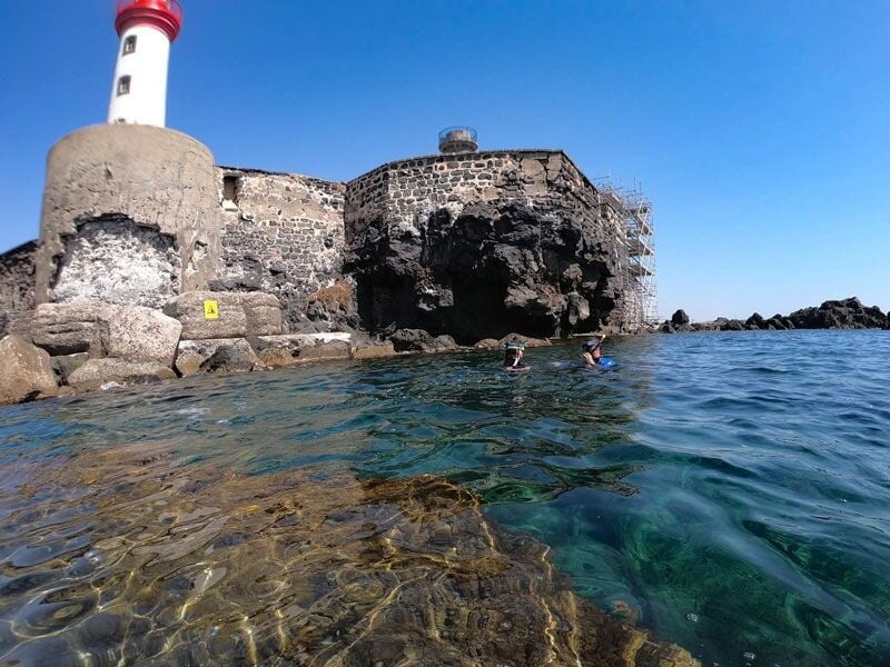 Phare rouge et blanc sur un fort en pierre au bord de l'eau claire avec deux personnes nageant à proximité.