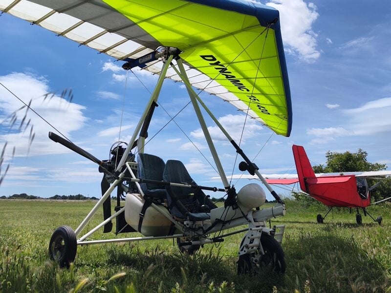 ULM stationné sur l'herbe avec un autre petit avion rouge en arrière-plan sous un ciel partiellement nuageux.