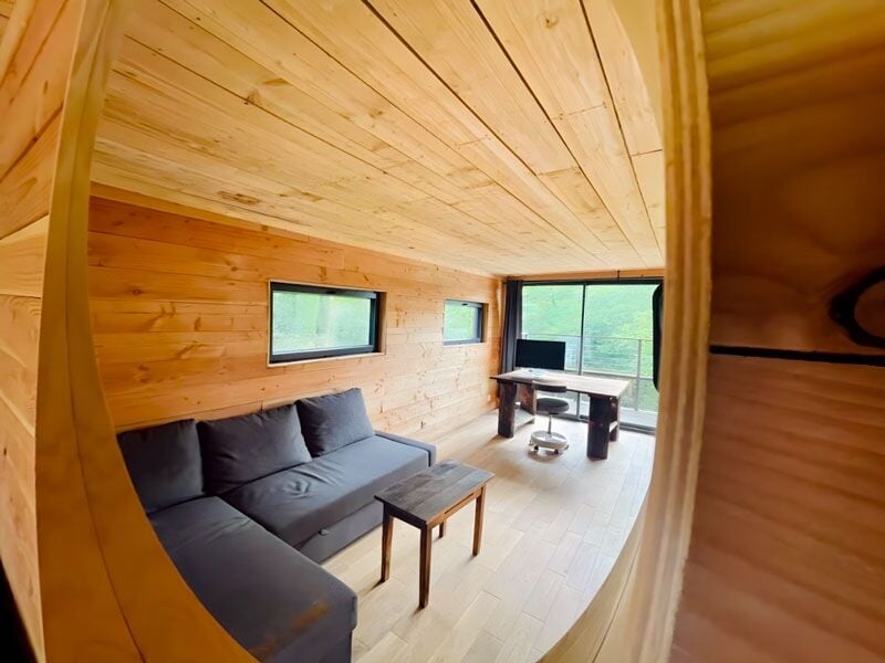 Salon en bois clair avec canapé noir, table basse et grande fenêtre donnant sur la nature.