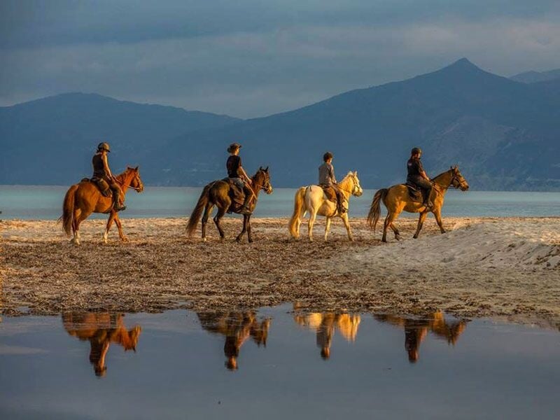 Quatre cavaliers sur la plage au bord de la mer avec montagnes et ciel nuageux en arrière-plan.
