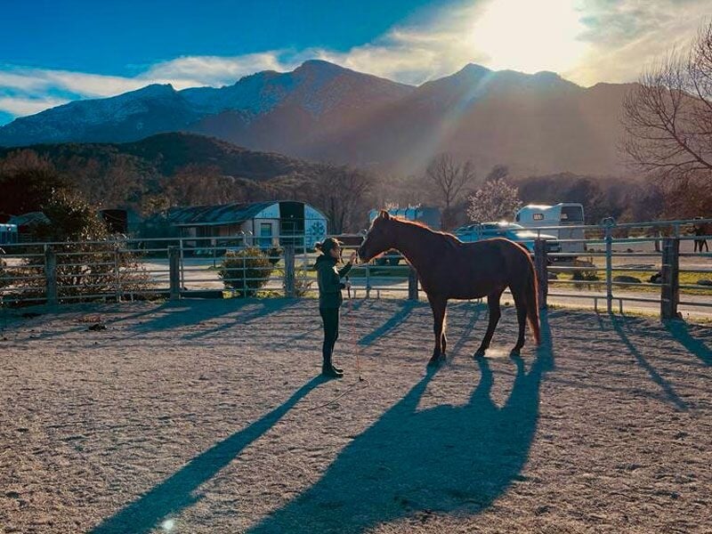 Personne tenant un cheval dans une écurie en plein air avec montagnes et soleil couchant.