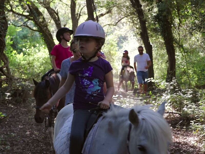 Enfants et adultes faisant une balade à cheval dans une forêt ensoleillée avec poussière.