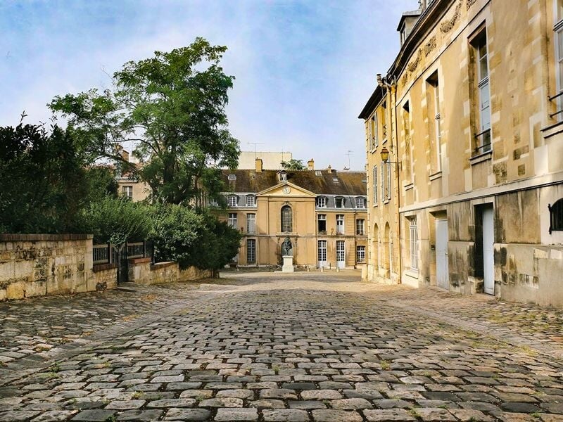 Rue pavée bordée de bâtiments anciens avec une statue au fond sous un ciel partiellement nuageux.