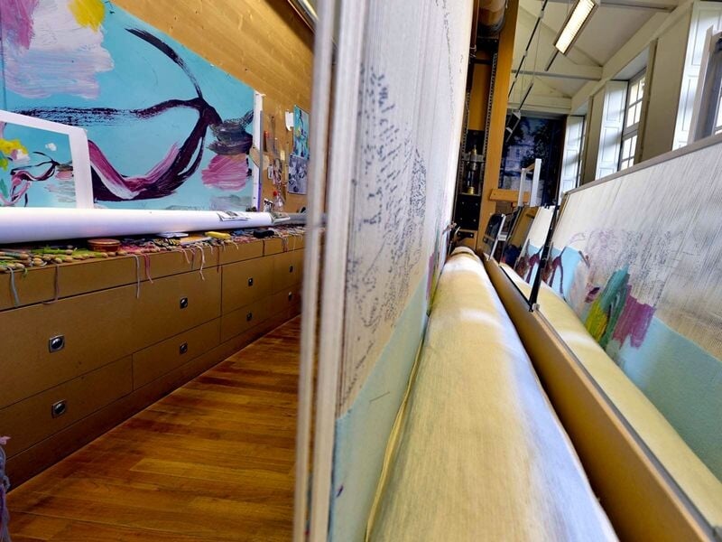 Vue rapprochée d'une tapisserie en cours de fabrication dans un atelier avec des peintures en arrière-plan.