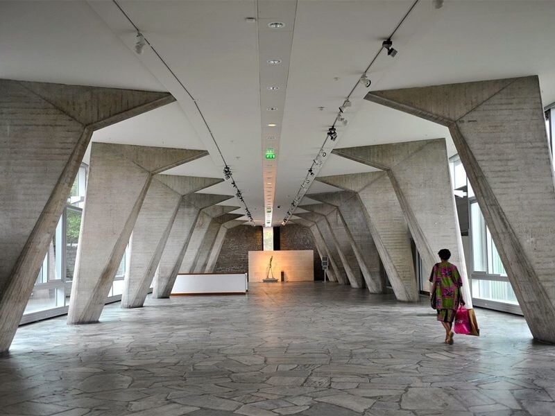 Couloir moderne avec piliers en béton inclinés et une personne marchant vers une lumière au fond de la pièce.