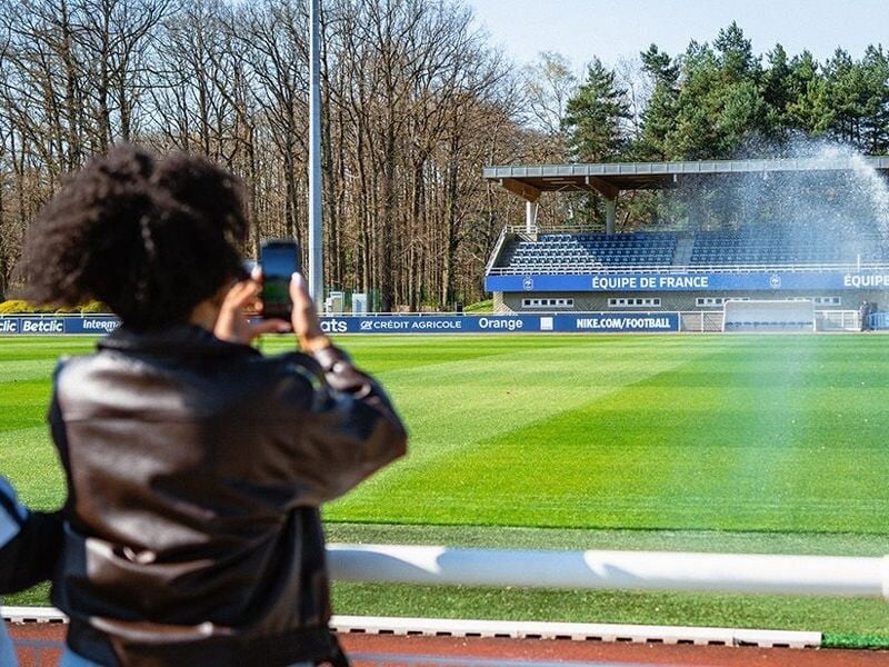 Personne prenant une photo du terrain de football avec les gradins et un arrosage automatique en arrière-plan.