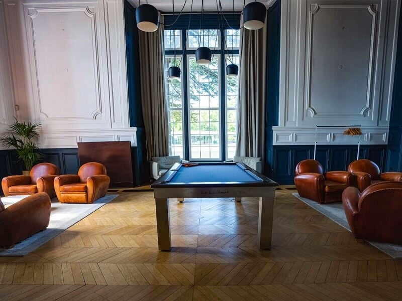 Salle élégante avec table de billard, fauteuils en cuir et grandes fenêtres laissant entrer la lumière.