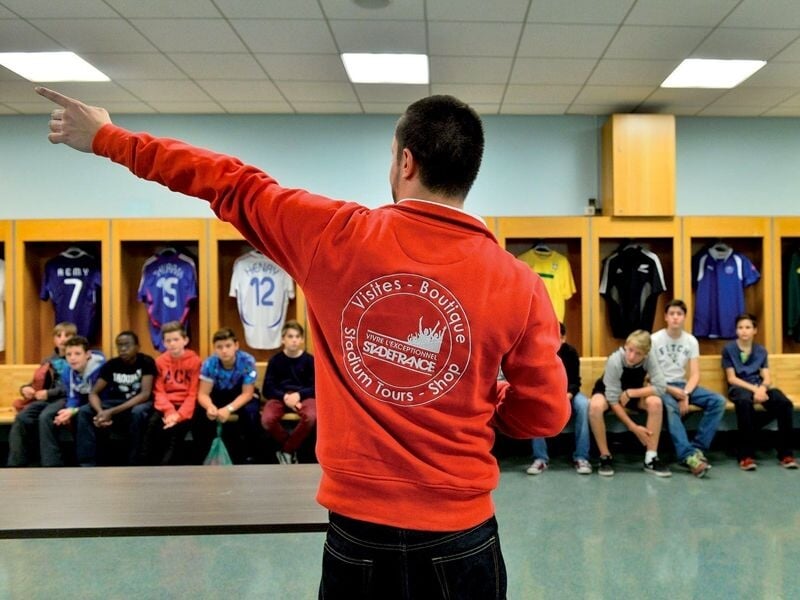 Un guide en sweat rouge montre quelque chose à un groupe d'enfants assis dans un vestiaire de football.