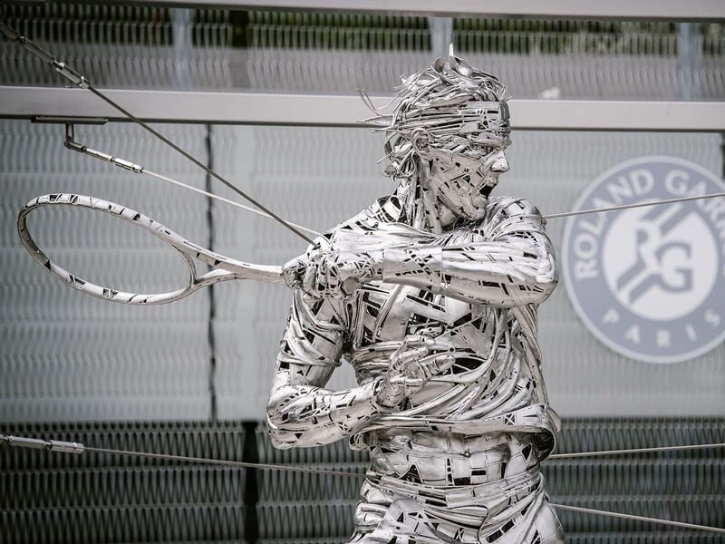 Sculpture métallique représentant un joueur de tennis en action avec une raquette, devant un mur avec logo Roland-Garros.