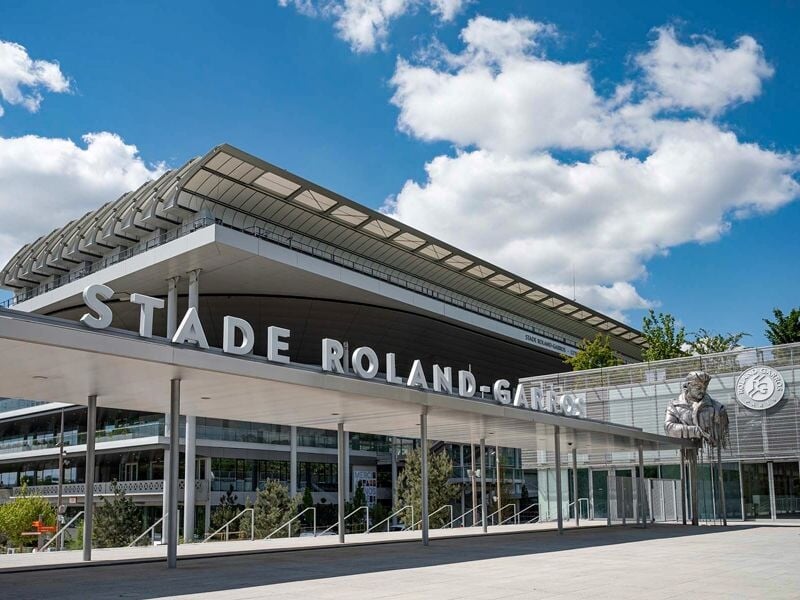 Façade extérieure du stade Roland-Garros avec son nom en grandes lettres blanches et un ciel bleu avec nuages.