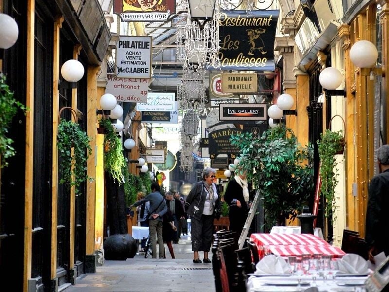 Passage urbain animé à Paris avec des enseignes, des plantes et des personnes marchant entre les boutiques.