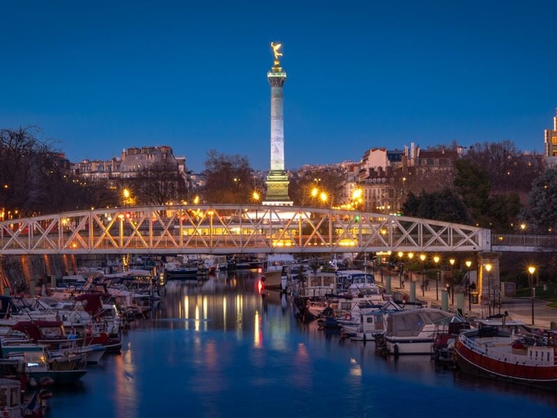 Vue nocturne d'un canal à Paris avec pont illuminé, bateaux amarrés et colonne de la Bastille.