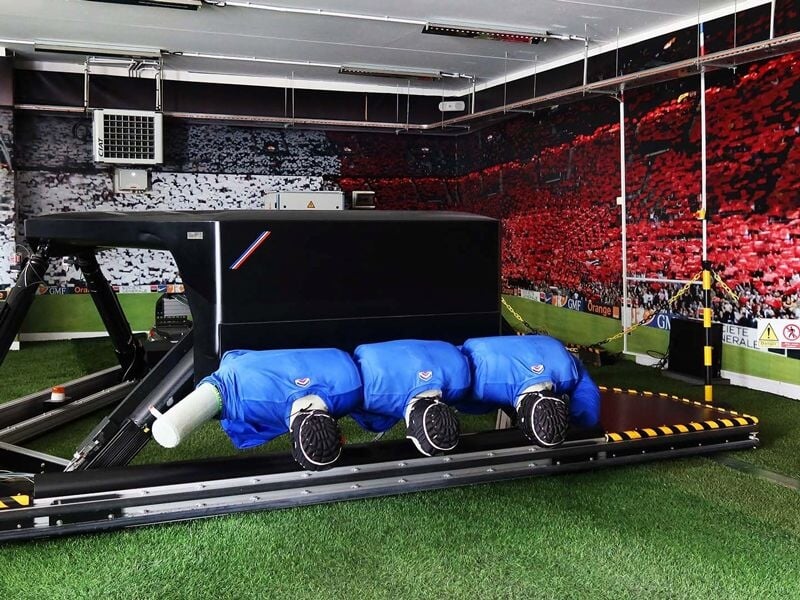 Salle d'entraînement de rugby avec mannequins bleus pour simulation de mêlée sur un sol en gazon synthétique.
