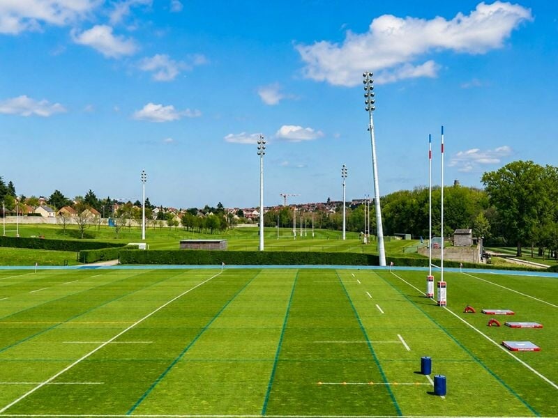 Terrain de rugby extérieur avec poteaux, pelouse bien entretenue et ciel bleu avec quelques nuages.