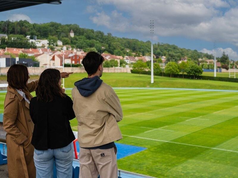 Trois personnes observant un grand terrain de rugby vert avec poteaux et éclairage en journée ensoleillée.