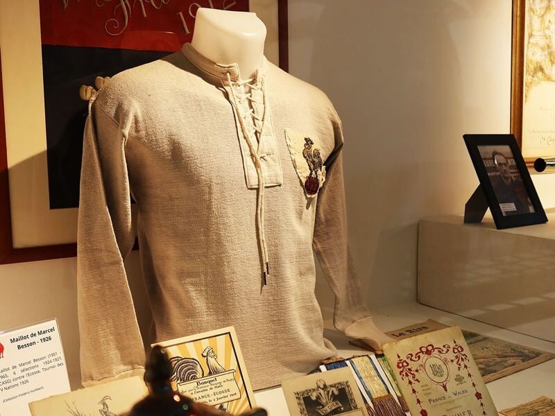 Vieux maillot de rugby exposé dans une vitrine avec documents et objets historiques autour.
