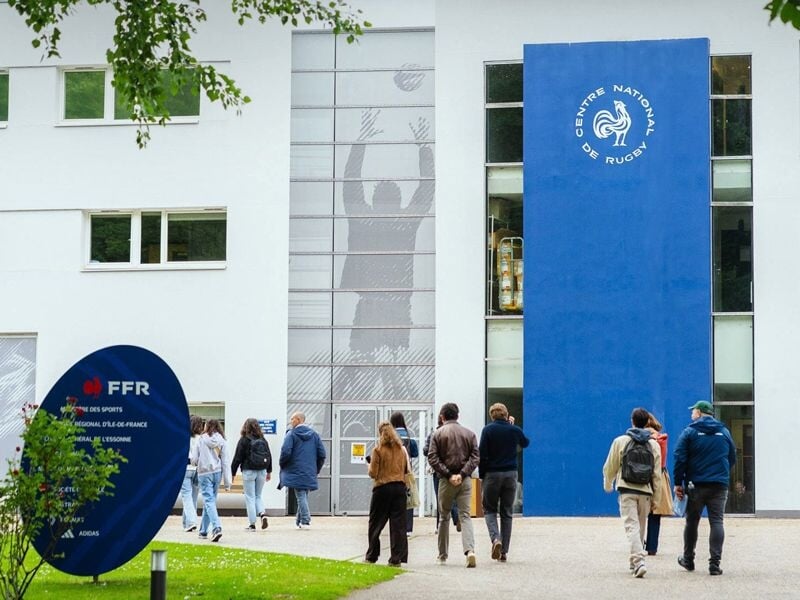 Entrée du Centre National de Rugby avec des visiteurs marchant vers le bâtiment blanc et panneaux bleus.