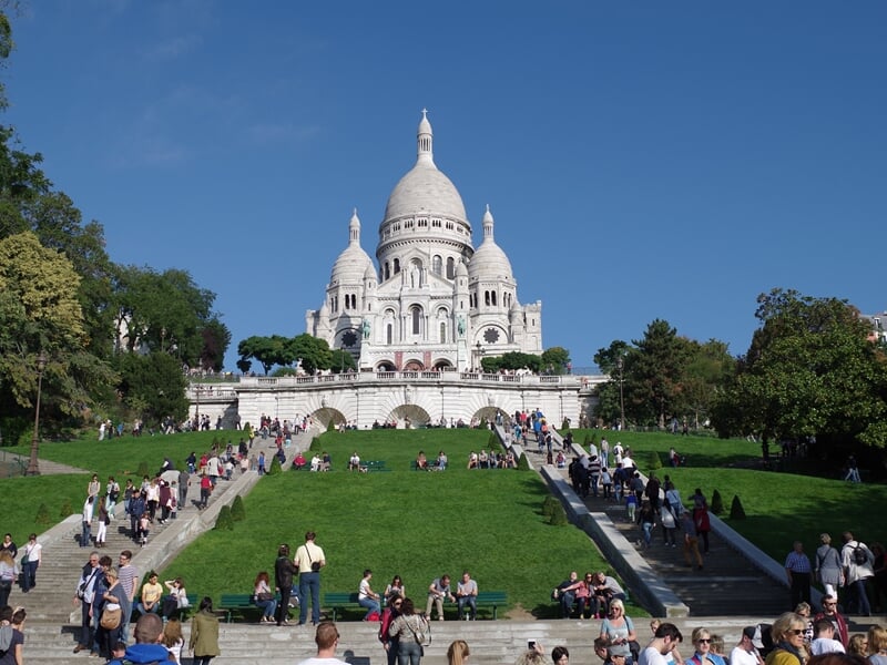 Foule de visiteurs assis et marchant sur les escaliers menant à la basilique du Sacré-Cœur sous un ciel bleu.