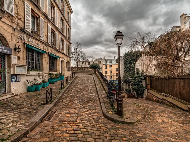 Rue pavée typique de Montmartre avec des bâtiments anciens, des plantes en pots et un lampadaire en fer forgé.