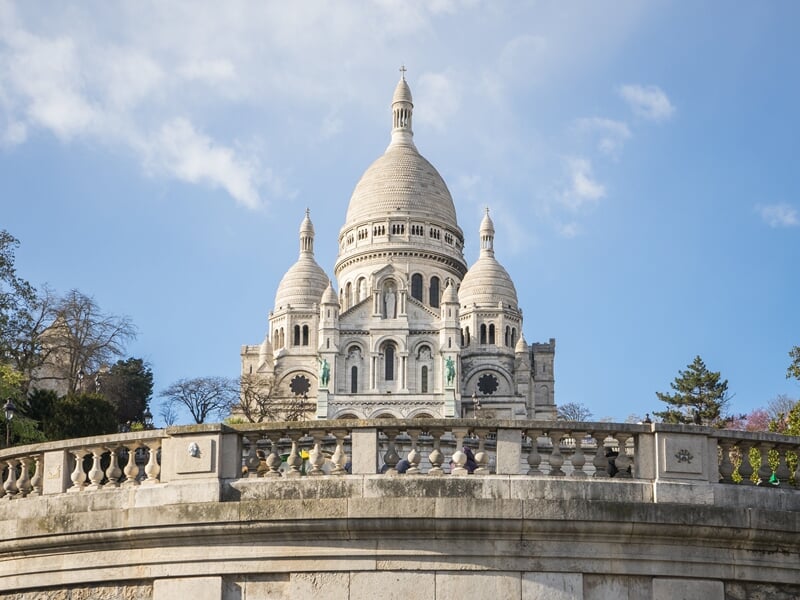 Vue frontale de la basilique du Sacré-Cœur avec un ciel bleu clair et une balustrade en pierre au premier plan.