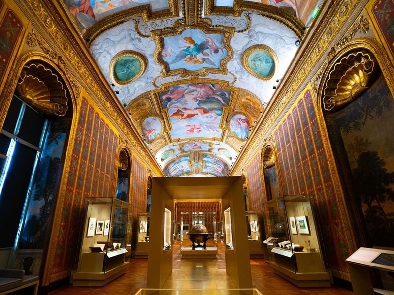 Grande salle richement décorée avec plafond peint et vitrines d'exposition dans une bibliothèque historique.