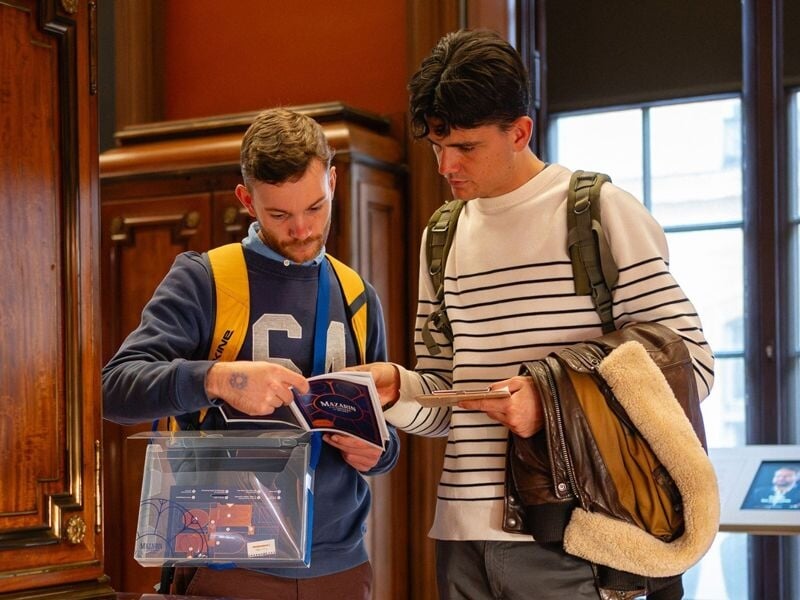 Deux hommes discutent en consultant un livret dans une salle en bois d'une bibliothèque ancienne.