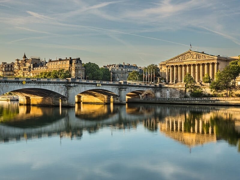 Vue du pont et des bâtiments historiques de Paris reflétés dans la Seine au coucher du soleil, ambiance paisible et dorée.