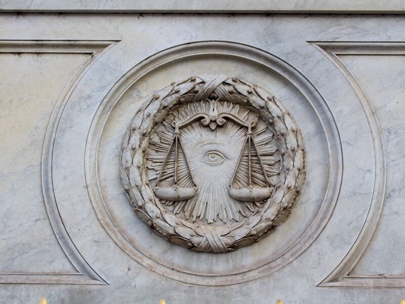 Détail sculpté en pierre représentant une balance et un œil entourés d'une couronne de feuilles sur un mur.