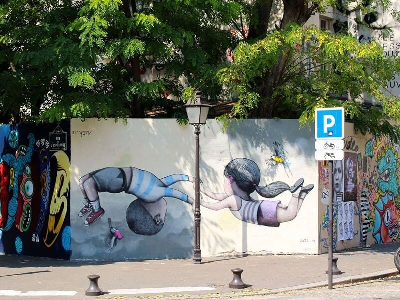 Fresque urbaine avec deux enfants jouant, un lampadaire au centre, arbres et graffitis colorés en arrière-plan.