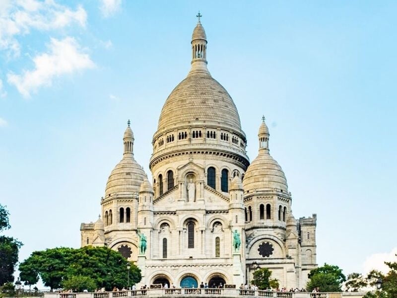 Vue frontale de la basilique du Sacré-Cœur de Montmartre sous un ciel bleu clair, entourée d'arbres verts.