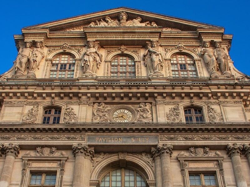 Façade ornée du Pavillon Sully au Palais du Louvre avec statues et horloge sous un ciel bleu clair.