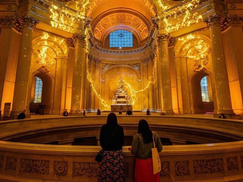Deux visiteurs admirant les projections dorées et lumineuses sur les murs et le dôme de l'Hôtel des Invalides.