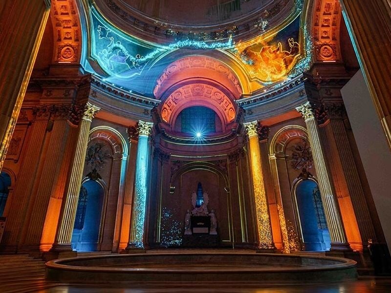 Vue intérieure de l'Hôtel des Invalides avec des projections colorées bleu, orange et jaune sur les colonnes et murs.