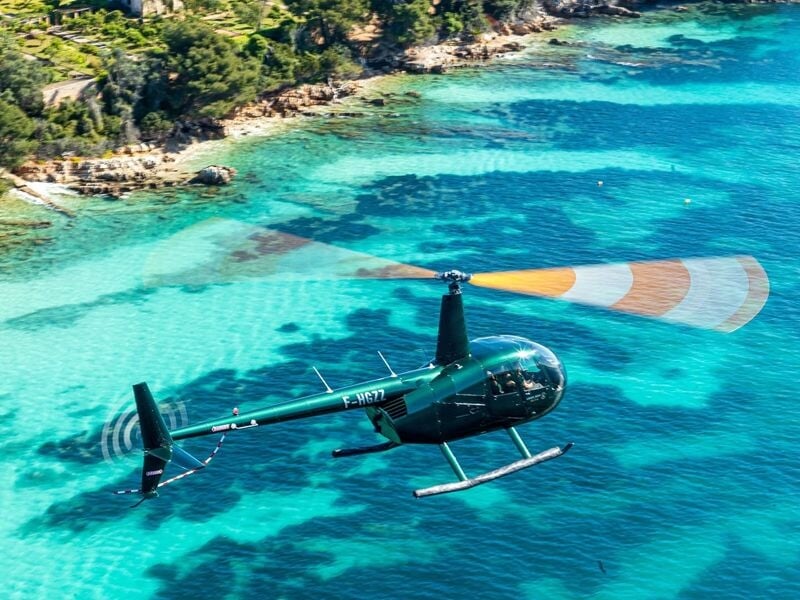 Hélicoptère vert volant au-dessus d'une eau turquoise claire près d'une côte rocheuse et végétalisée.