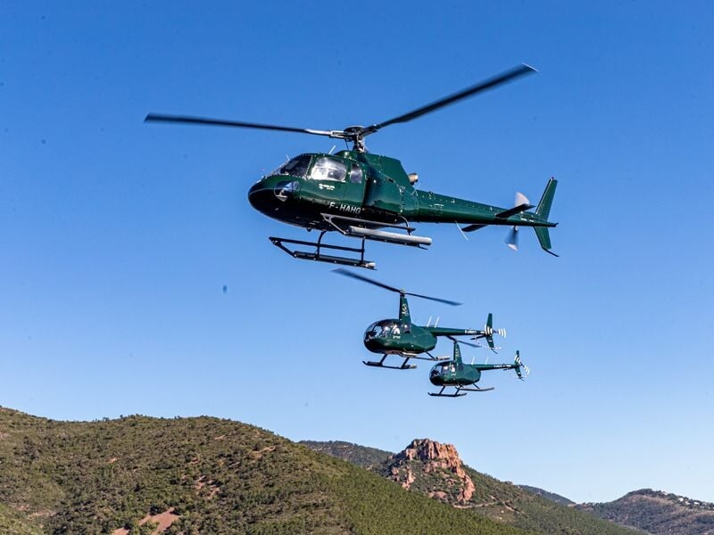 Trois hélicoptères verts volant en formation au-dessus d'un paysage montagneux sous un ciel bleu.