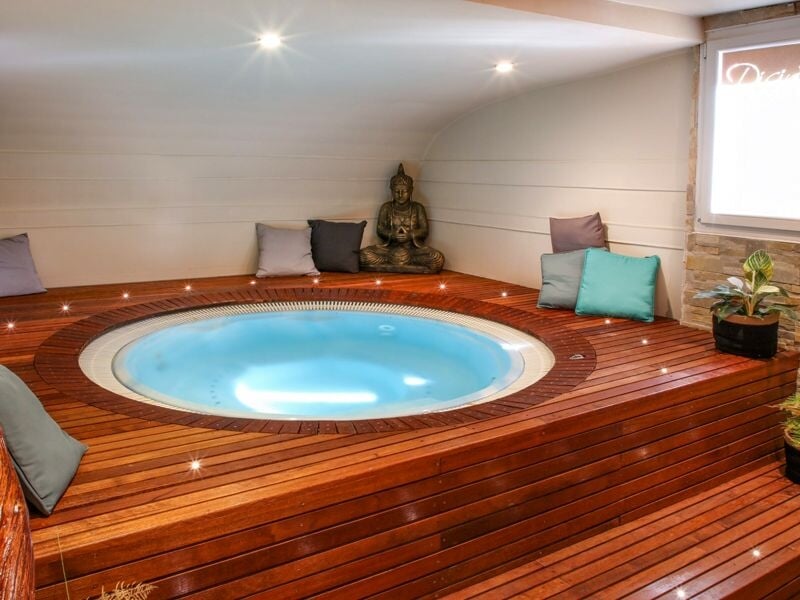 Un jacuzzi rond encastré dans un sol en bois avec des coussins autour, dans une pièce lumineuse et accueillante.