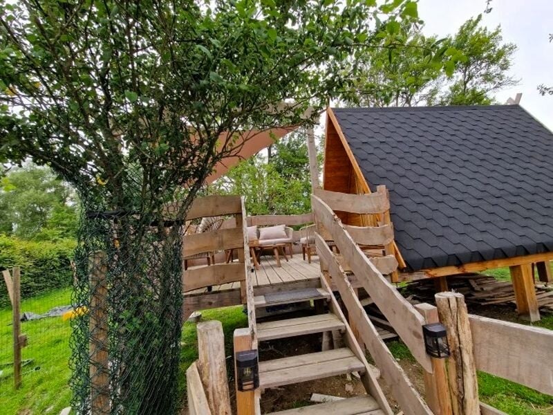 Escalier en bois menant à une terrasse avec une petite cabane en bois au toit noir dans un jardin verdoyant.